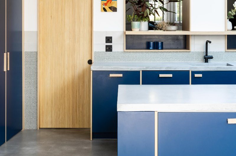 Blue kitchen with wood handles, concrete-effect surfaces, a sliding wood door, and black accents such as the tap and sockets.