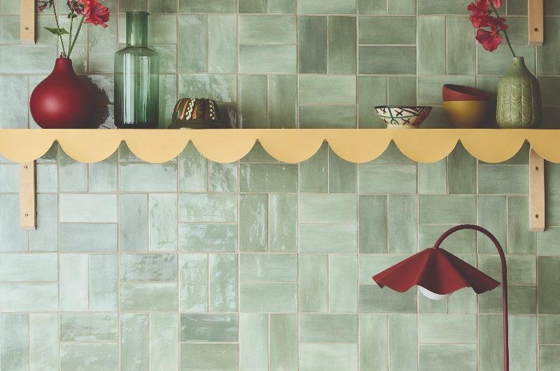 Kitchen with sage green tiles, scalloped shelving, and burgundy decor.