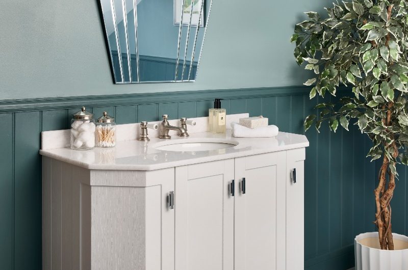 A classic bathroom with white furniture, green wall paint and panelling and an art deco wall mirror.