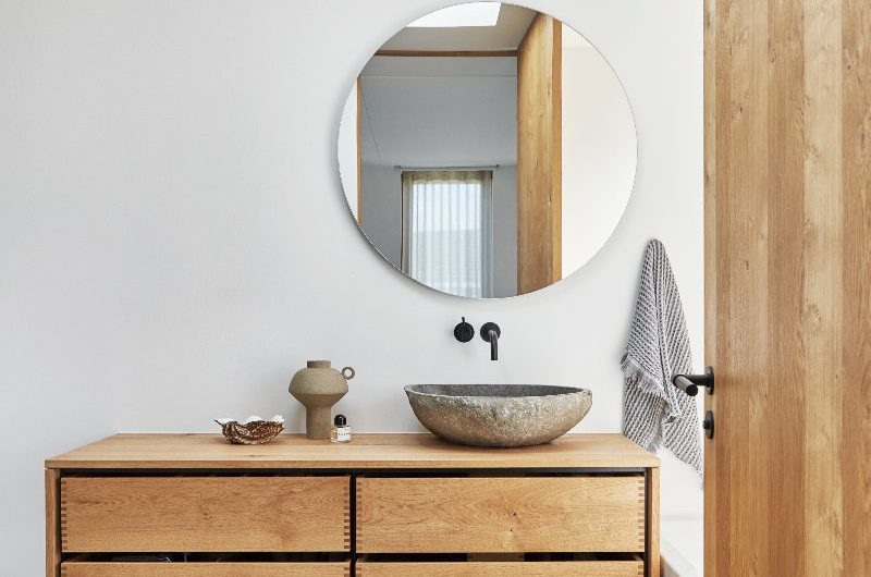A modern minimalist scheme with wood furniture, stone basin, circular mirror and Scandi decor.