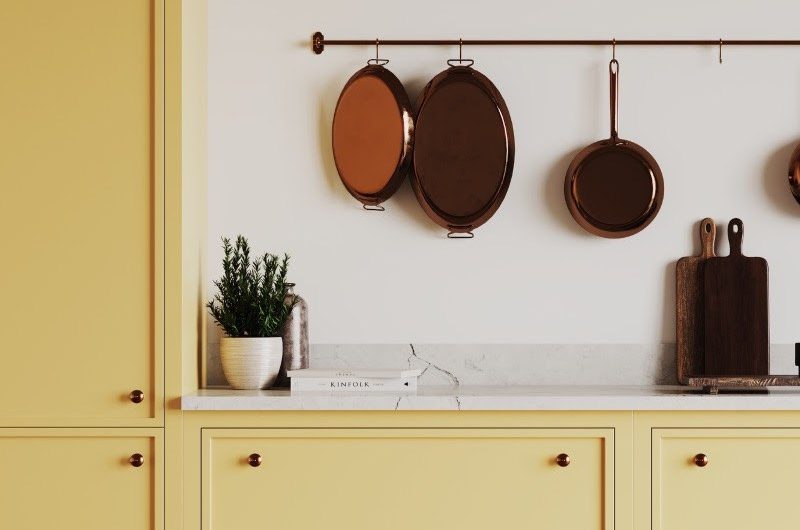 Slim Shaker kitchen cabinetry painted in a light yellow with veiny worktops and a brass rail holding pans.