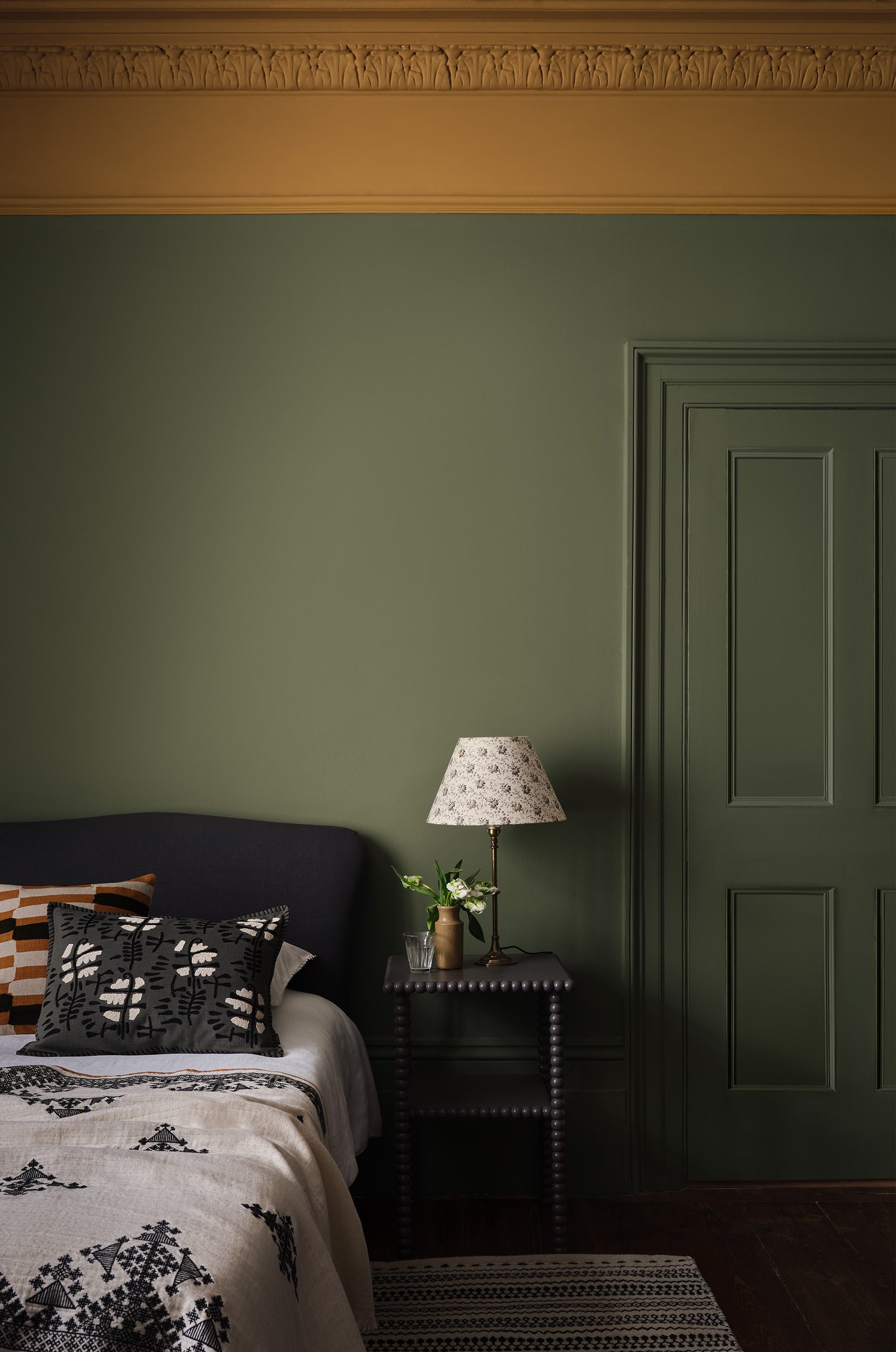 Dark green bedroom with a simple bed, minimalist bedding, bobbin side table, and mustard yellow paint on the cornicing.