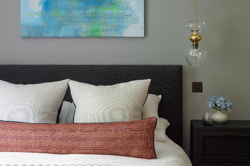 Green bedroom with a dark grey upholstered frame, neutral, tactile bed linen and cushions, and a dark rattan side table.