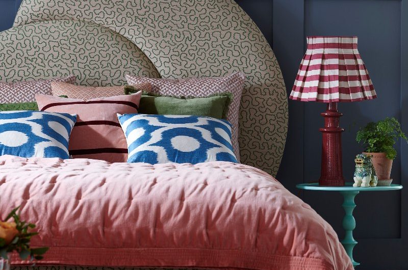 A bold scheme with a patterned headboard against dark blue wall panelling and floral wallpaper, with colourful bedding and a teal side table.