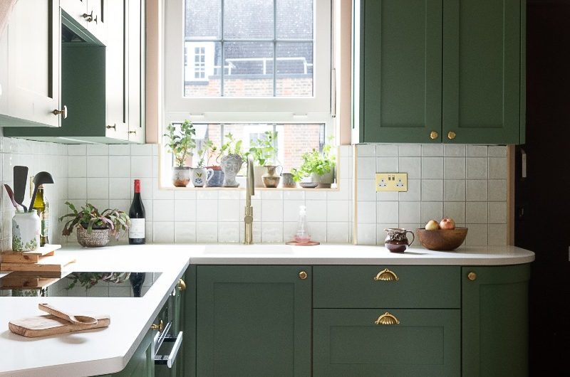 Green l-shaped kitchen in a small space, with white splashback tiles and worktops, hexagon terracotta flooring and gold accents.