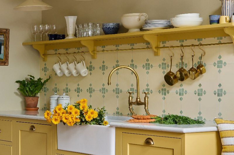 Yellow kitchen with classic cabinetry, a butler sink, brass tap, patterned tiles, and an open shelf for storage.