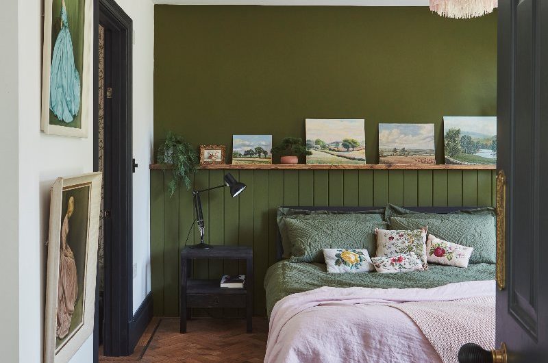 Bedroom design with a deep olive green panelled wall, floral cushions and paintings on the wall.