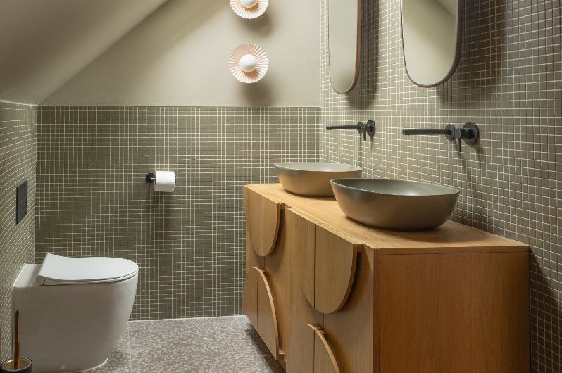 A retro modern bathroom with mosaic green tiles, terrazzo flooring, mid-century furniture, countertop basins and wall-mounted taps.