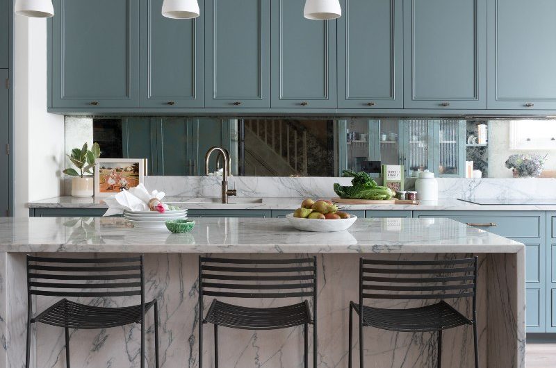 A blue kitchen with a marble island and breakfast bar with industrial touches such as metal chairs and lighting.