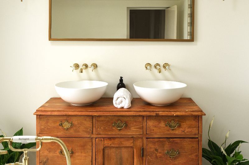A vintage vanity unit with classic hardware, two countertop basins, gold brassware and an antique mirror with matching wall lights next to it, in a spa-like bathroom.