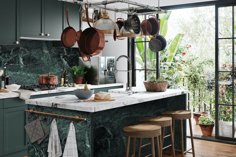 A green kitchen with a marble island with pots and pans hanging from the ceiling.