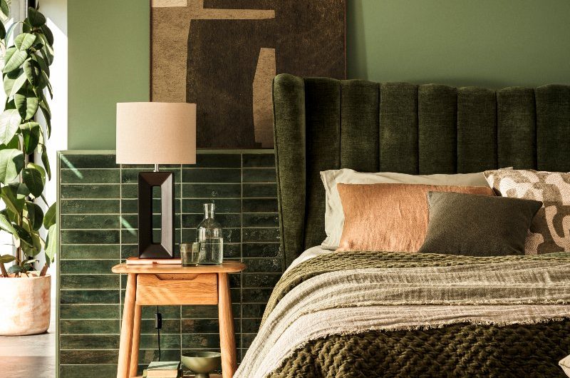 Green bedroom design with a velvet headboard, green tiles behind it, wood flooring and furniture, and house plants.