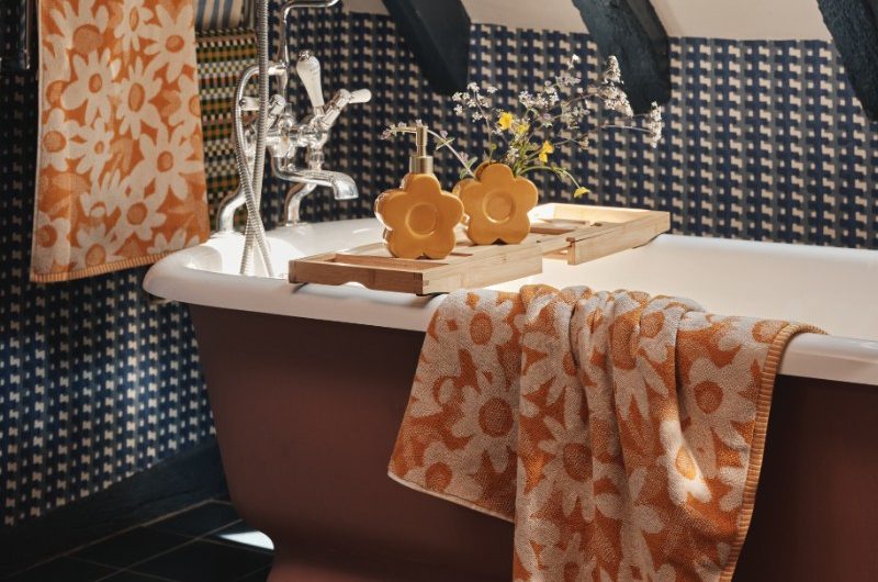 Country bathroom design with blue beams, patterned wallpaper, a burgundy tub with a wood bath tray, chrome bath filler and floral towels in white and yellow.