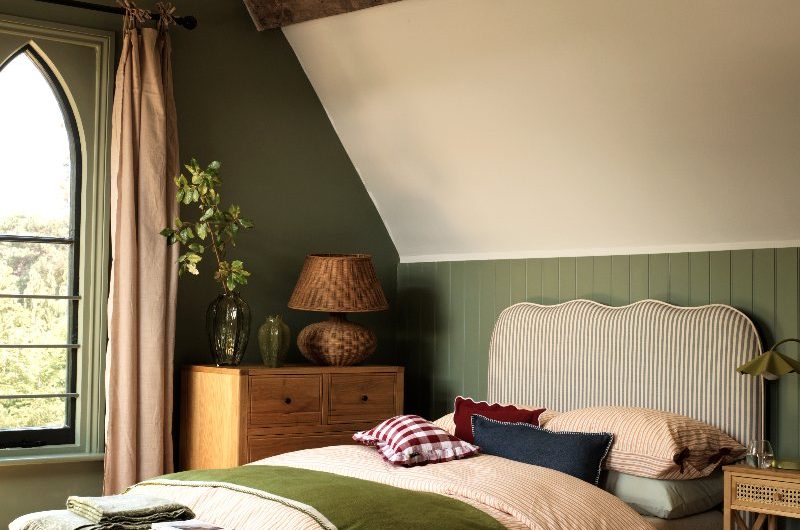 Country autumn-themed bedroom with a striped headboard, grey ottoman, wood furniture, green wall panelling, and wood beams and flooring.