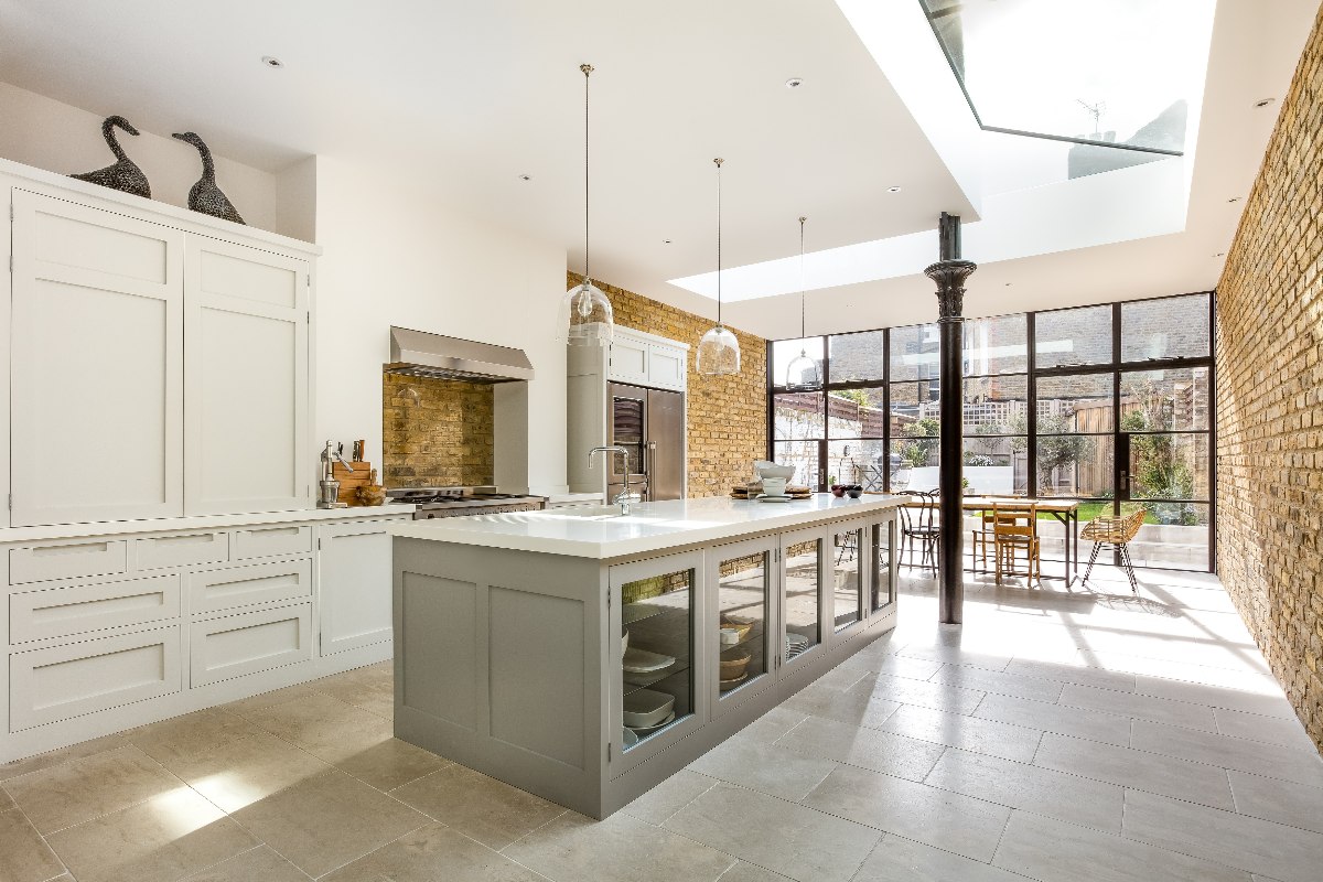A galley kitchen design with a large island with glass door fronts, white surfaces, stone flooring, exposed brick walls and a skylight.