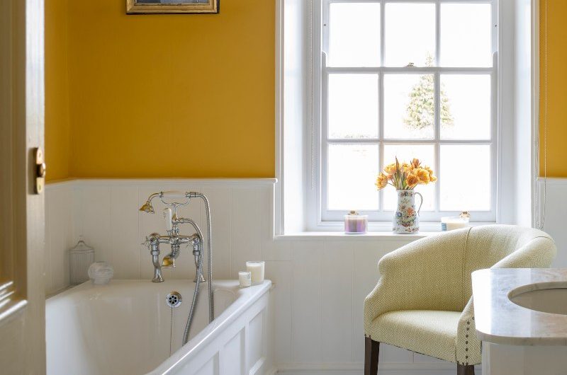 A classic bathroom with white wall panelling on the lower half of the wall, yellow paint on the upper half, a fitted tub, timeless furniture and patterned window roller blind.