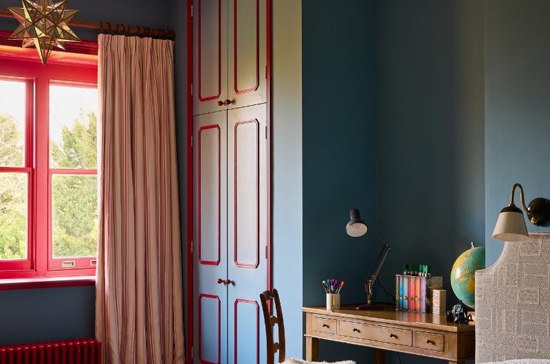 Kids' bedroom with blue wall paint, pops of red on the window, radiator and wardrobe, and a compact desk area.
