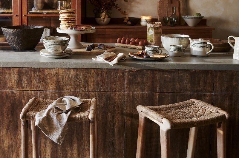 Rustic, autumnal kitchen design with plaster on the walls, a wood island with a concrete worktop, rattan bar stools, and white crockery.