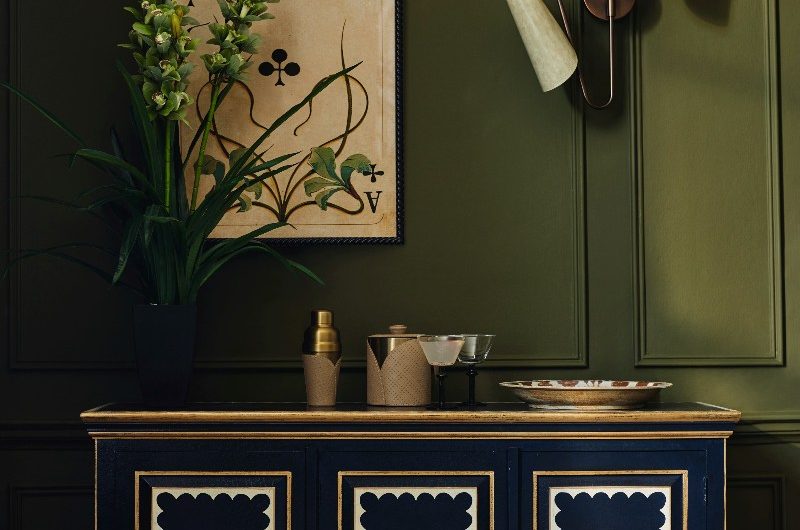 A compact bar area with a handcrafted, navy sideboard, green wall panelling, and herringbone flooring. 