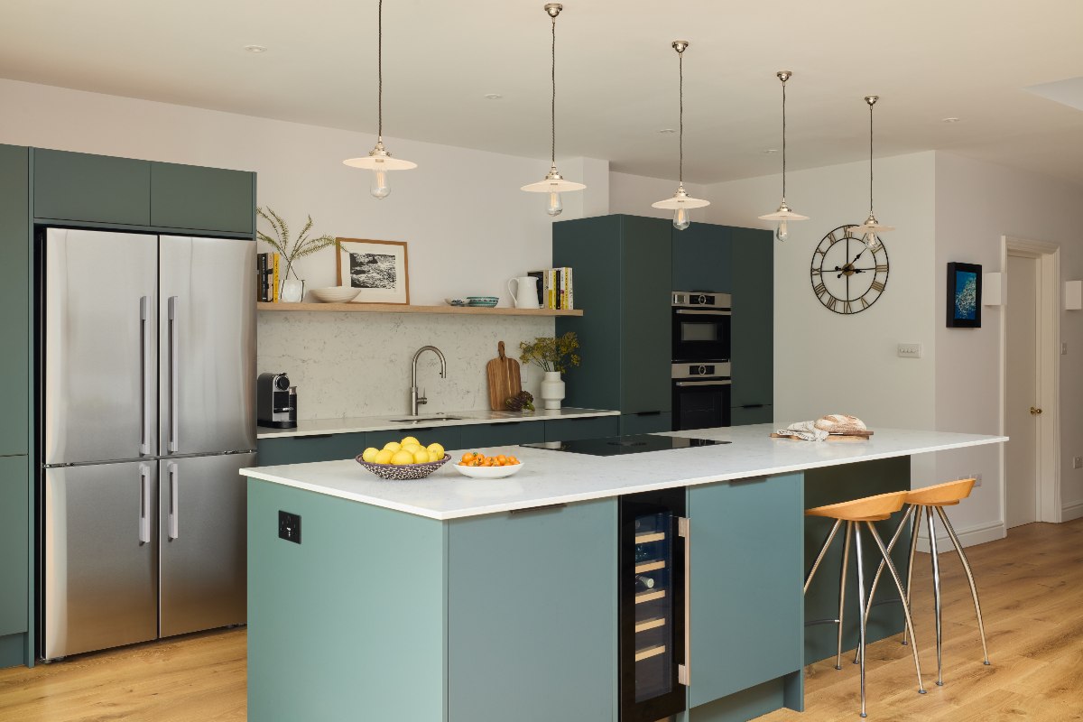 Handleless green kitchen with white worktop, breakfast bar with two stools, and built-in appliances.