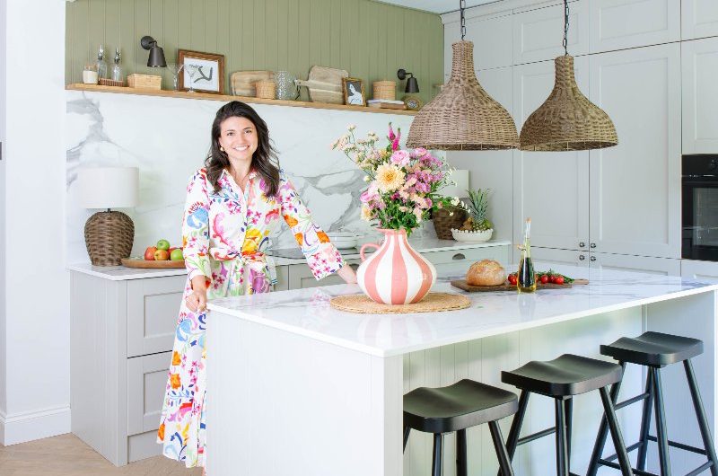 Homeowner Dot posing in her stunning kitchen makeover which includes neutral cabinetry, and island, and sage green panelling.