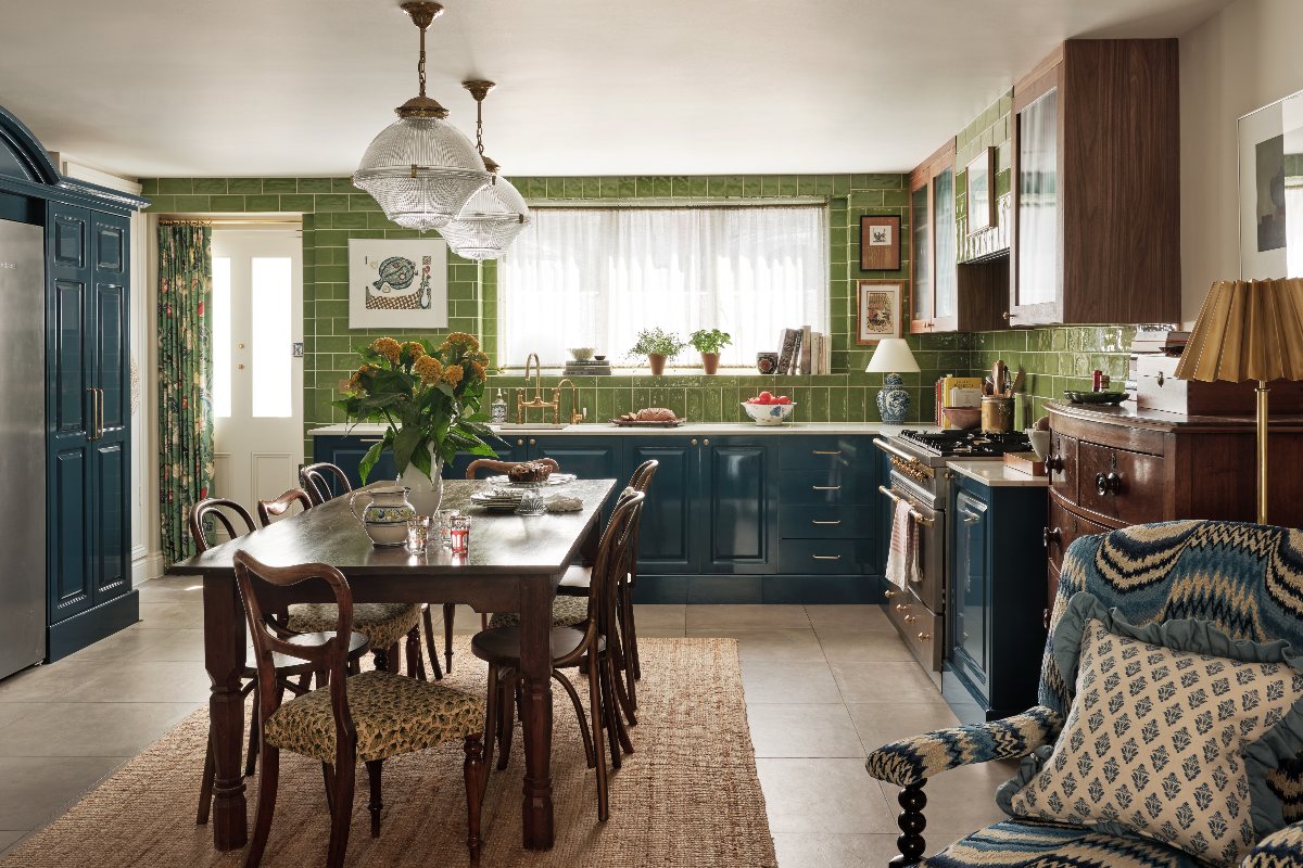 L-shaped green kitchen with green tiles, wood cabinetry and a dining table used as an island in the centre of the room.