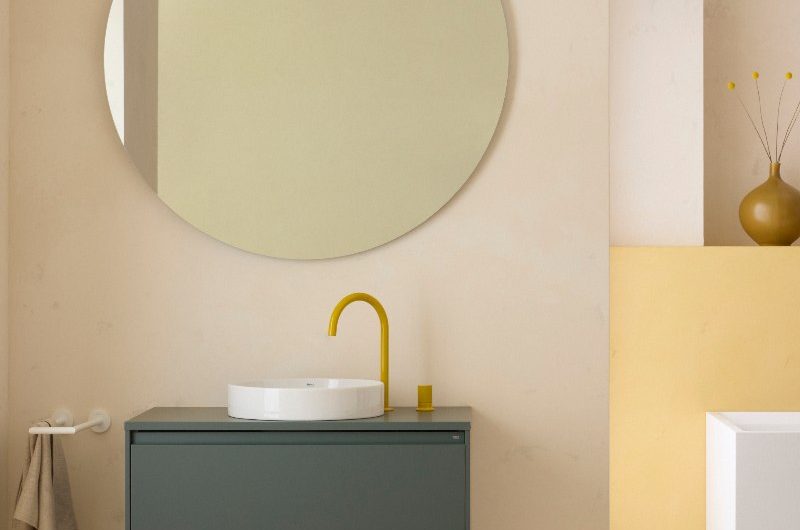 A neutral bathroom with a wall-mounted dark green vanity, compact countertop basin, yellow tap, and a grand circular mirror above it.