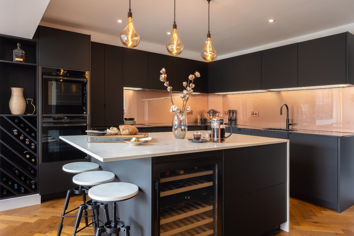 Dark handleless design with copper splashbacks, a central unit with a drinks fridge and seating, and industrial lighting above it.