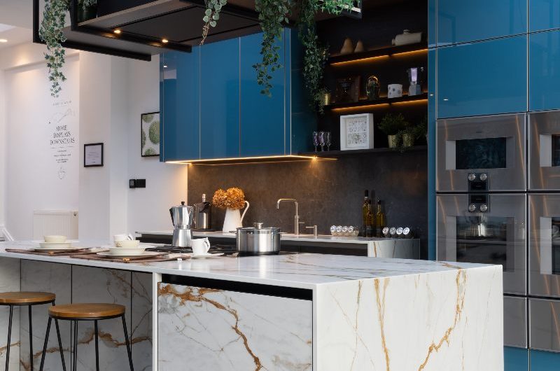 Blue handleless cabinetry with built-in appliances, a stone-clad island with storage and seating and a ceiling extractor hood.