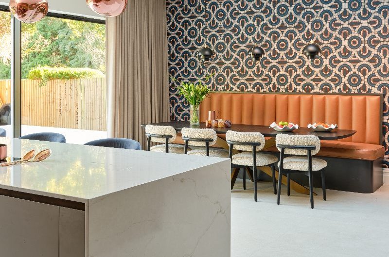 A retro, 70s dining area with statement wallpaper, a banquette seat, large table, and chairs under a skylight.
