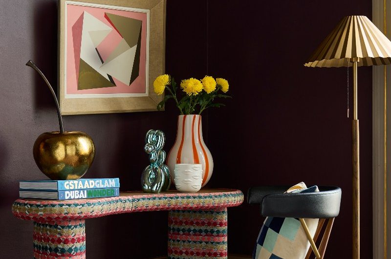 A colourful sideboard in a wine-coloured room, with a leather chair, brass floor lamp and patterned rug.