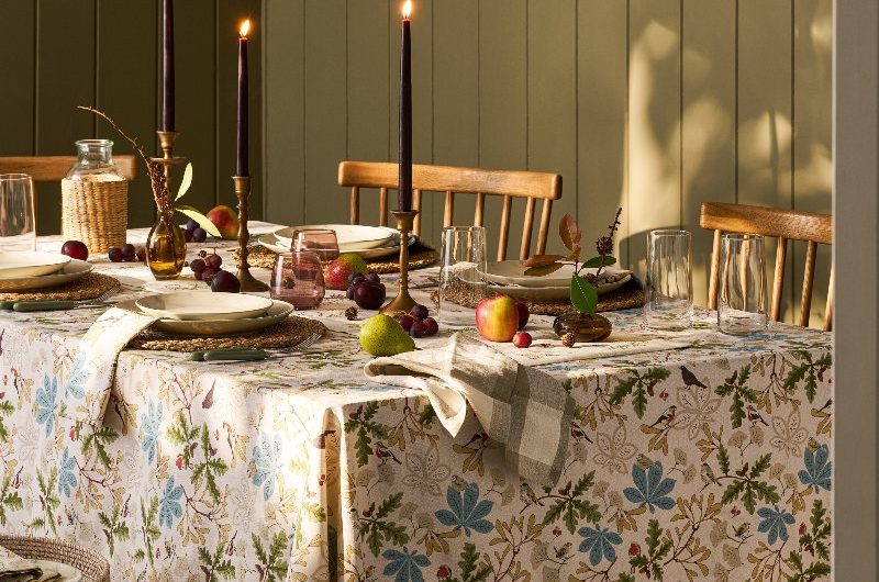 Autumn dining room with green wall panelling, and a table decorated with a patterned tablecloth, seasonal harvest, black candle sticks, wood chairs and rattan pendants.