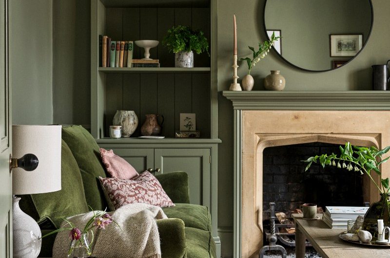 Autumn snug drenched in green with a matching velvet sofa, bookcase, wooden coffee and side tables and a cosy fireaplace.