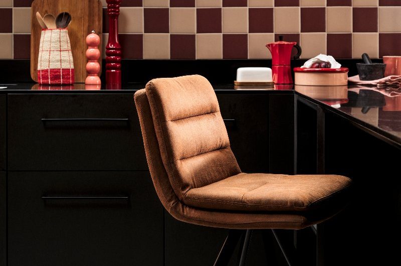 L-shaped kitchen with dark cabinets, cream and burgundy checkerboard tiles, velvet bar stool, and a wood shelf.