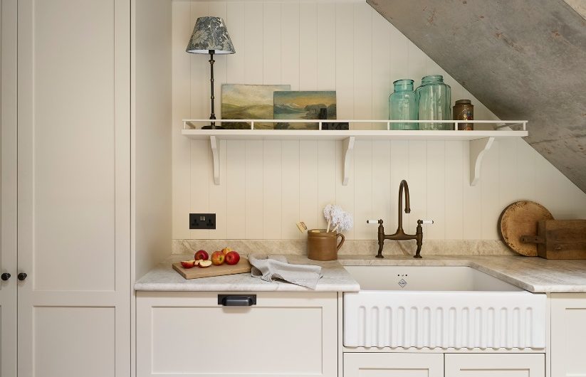 A compact kitchen's sink zone fitted under the stairs, with cream cabinetry, neutral worksufraces, a fluted sink, and brass tao.