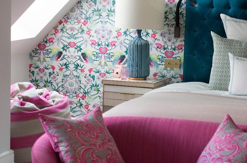 A colourful bedroom design with pink and blue wallpaper, velvet furnishings and patterned cushions, flooded with light through a skylight.