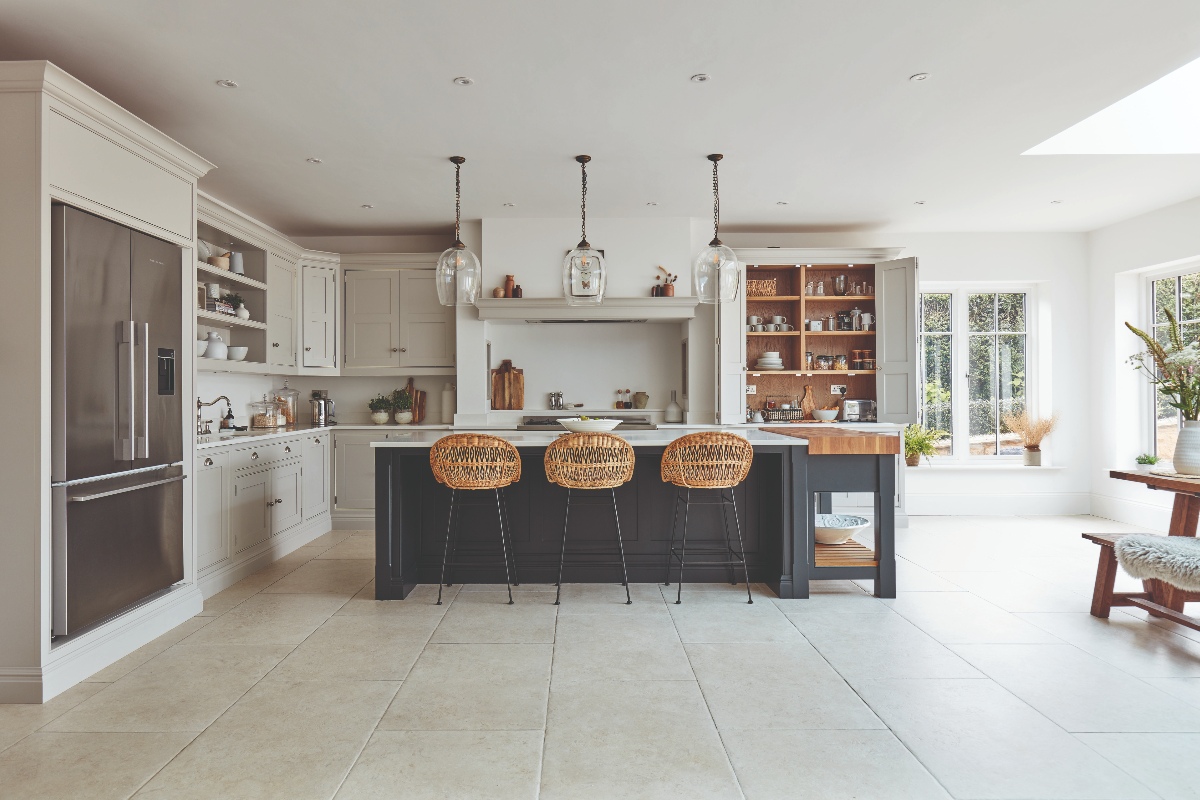 L-shaped light grey cabinetry with a breakfast cupboard, range cooker area, sink and tap zone and built-in fridge, and a central island with seating and a wood butcher block.