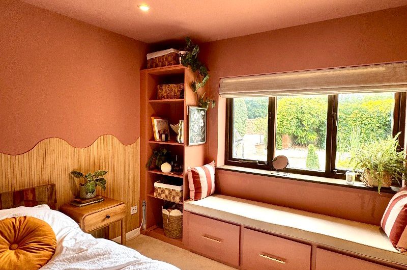 A peach pink bedroom with a fitted bench, open shelving, wavy wood panelling, and bed frame.