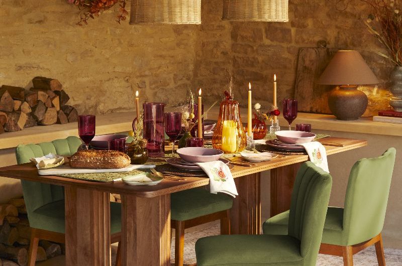 An autumnal table scheme with a wood table, green velvet chairs, burgundy glassware, and lit-up candlesticks. 