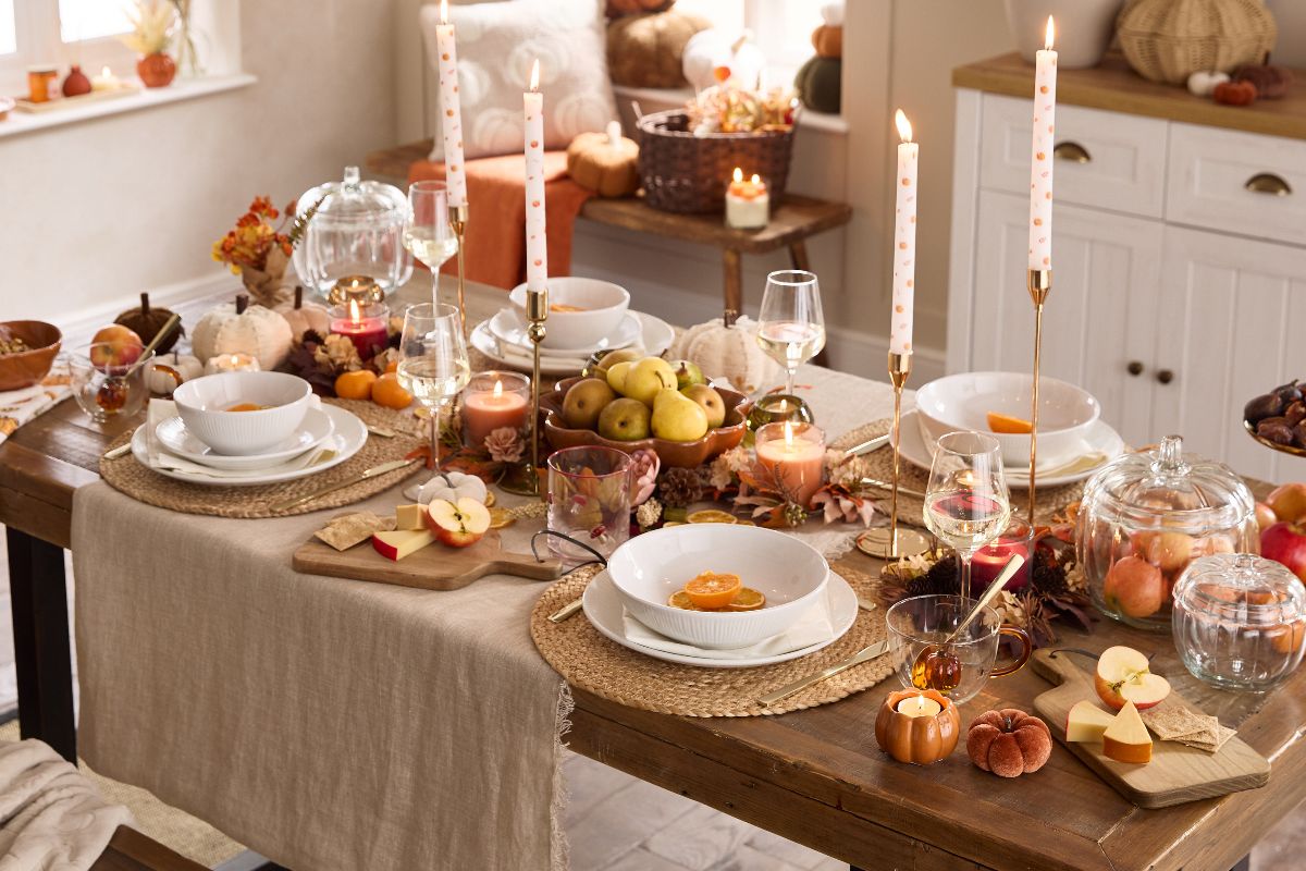 A classic autumn tablescape with pumpkin-shaped jars, and candle holders, white tableware, glass wine glasses and wooden boards.