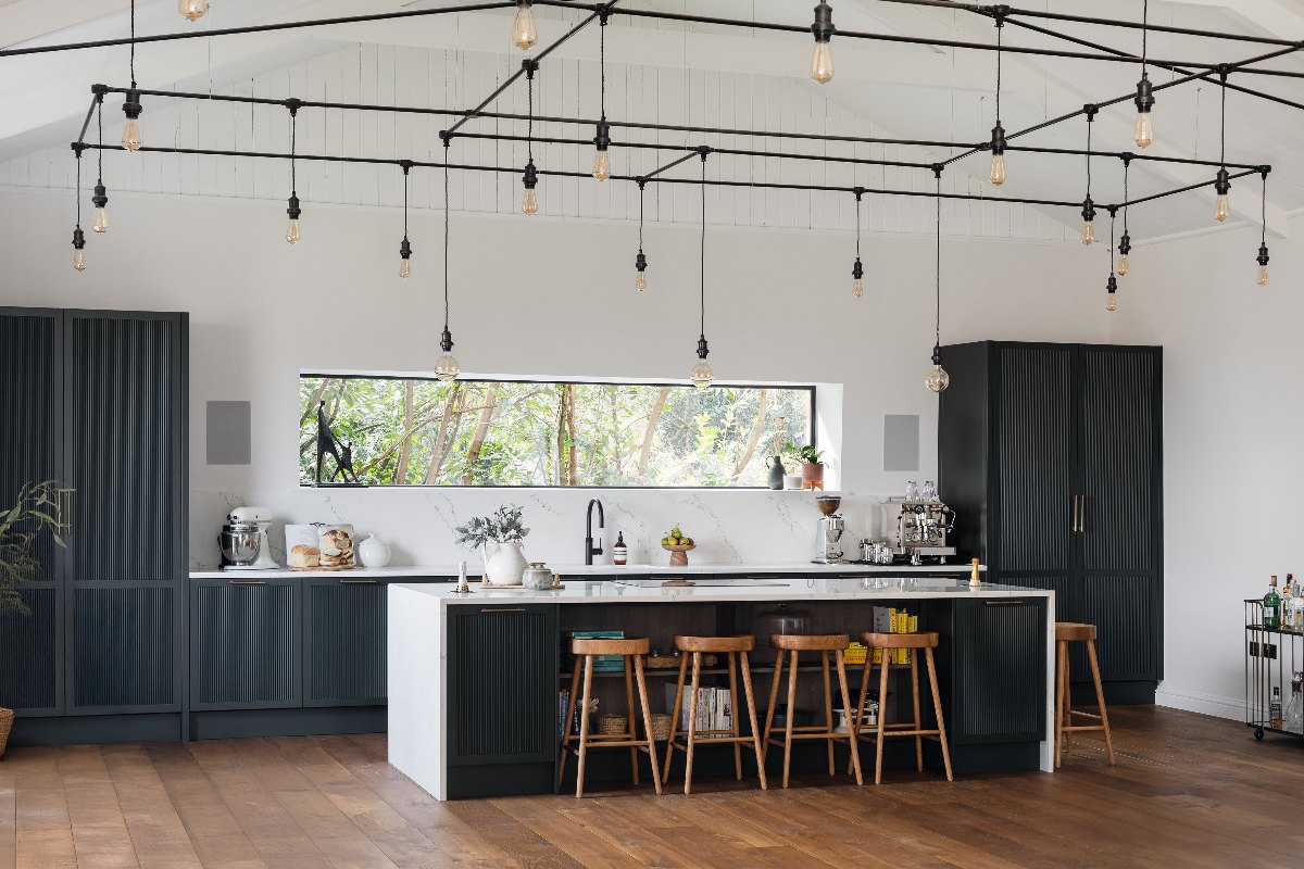 A dark green kitchen in a converted barn with statement lighting, fluted cabinetry and a central island.