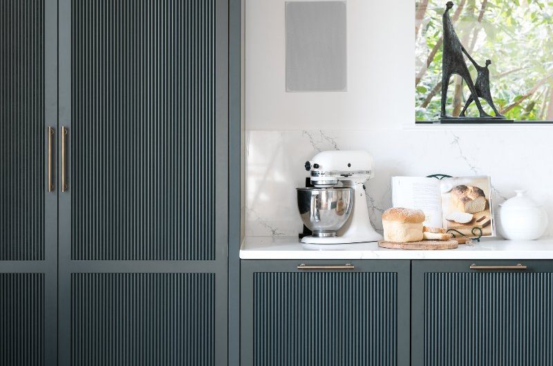 Tall dark green kitchen cabinets with bespoke fluting, sleek handles, with wood flooring and white work surfaces.