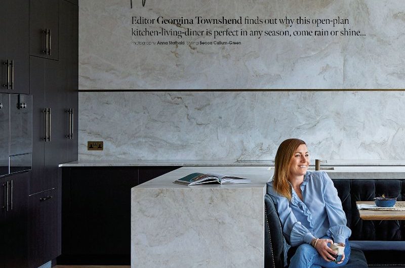 Homeowner sitting her banquette in an open-plan scheme with statement marble surfaces and dark cabinetry.
