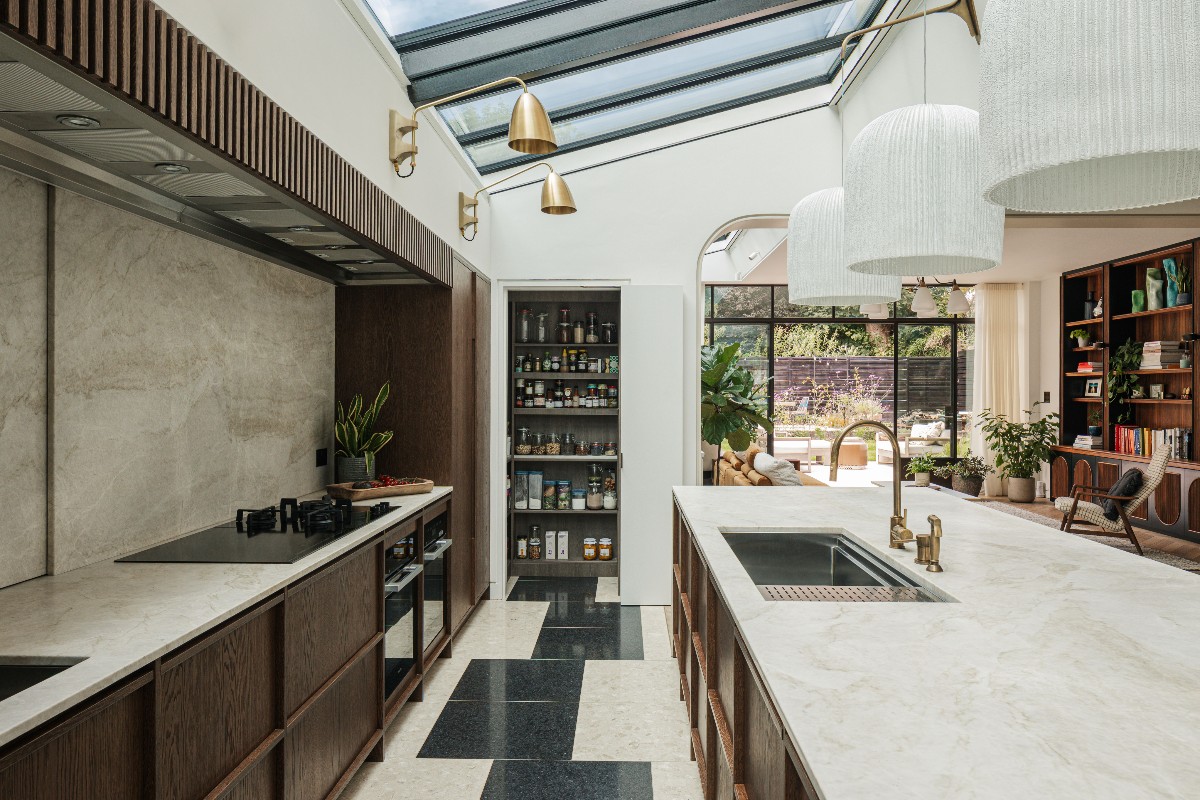 A cooking space with wood cabinetry, veined surfaces, concealed pantry, a sink and tap, and sleek, black appliances.