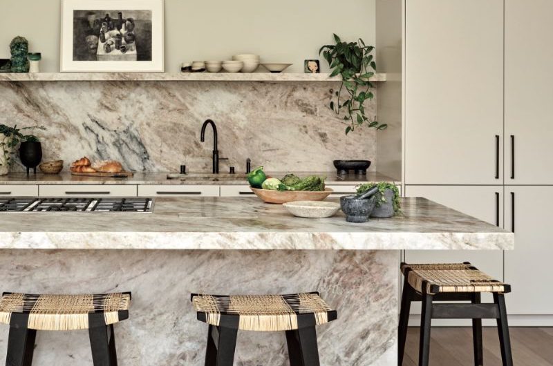 A closeup at a stone kitchen island, with sleek cream cabinetry, stone splashback and matching open shelf and natural materials.