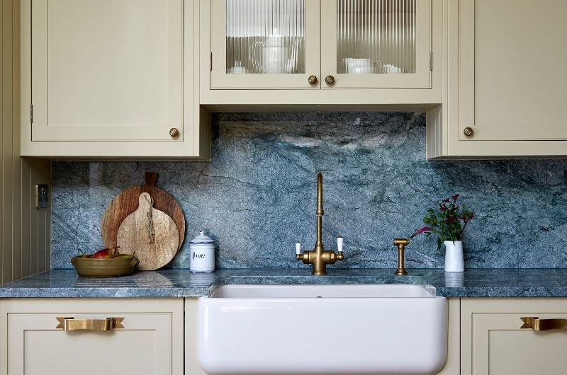 Sink zone with stone worktop and splashback, glazed cupboard and brass hardware.