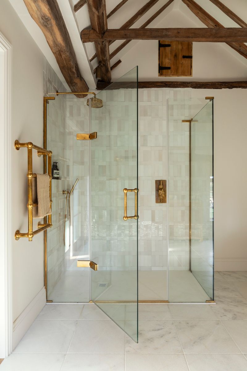 A shower design in a country-style bathroom with a gold framed glass enclosure, white glossy tiles, and gold brassware.