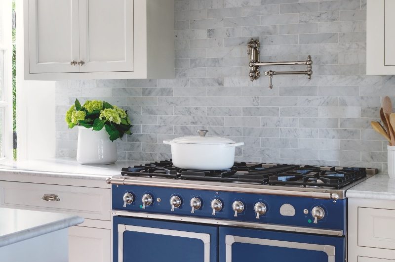 White kitchen with a statement blue range cooker, chrome pot filler tap, and marble tiles.