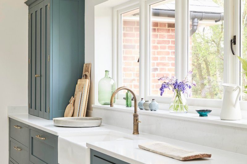 A run of cabinets with minimalist gold handles, white worktops, butler sink, a brass tap, which enjoys views of the garden.