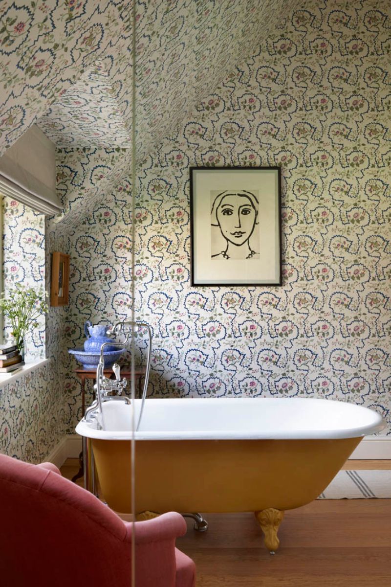 Yellow painted tub with chrome brassware, with quirky wallpaper, art and a pink armchair in the corner.
